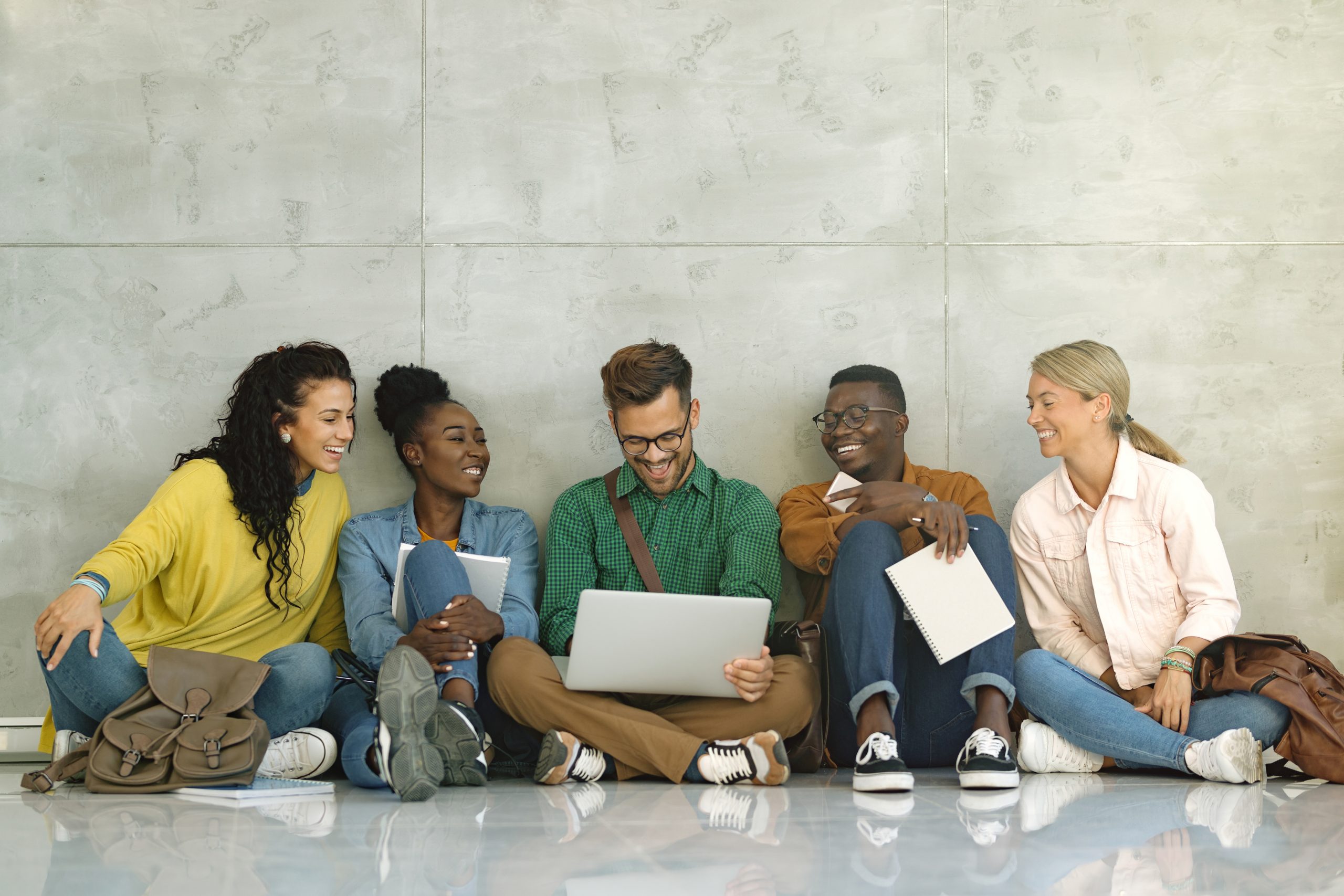 happy-university-students-using-laptop-while-sitting-hallway-1-scaled
